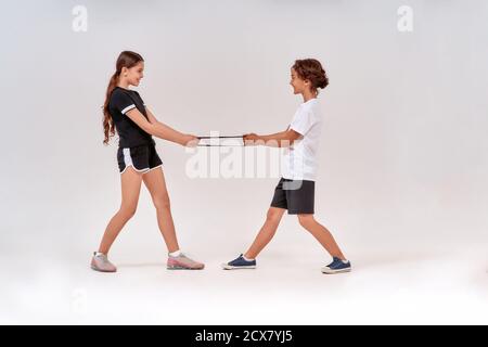 Foto a tutta lunghezza di felice ragazzo e ragazza adolescenti che si divertono mentre si esercita con la banda di resistenza, in piedi isolati su sfondo grigio Foto Stock