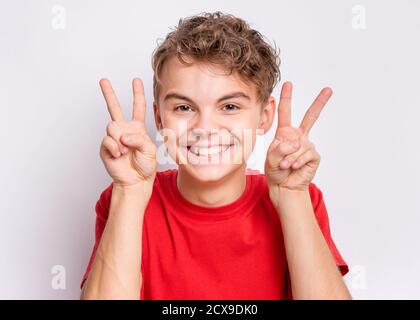 Ritratto teen boy facendo gesto della Vittoria, su sfondo grigio. Adolescente caucasico sorridente e mostrando segno di vittoria. Felice bambino che fa la vittoria simbolo. Foto Stock