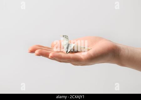 Mano femminile con uccello origami fatto di banconota da dollaro sopra sfondo chiaro Foto Stock