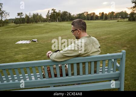 Vista posteriore di un giovane bel ragazzo seduto sul panca in legno nel parco e con tablet digitale Foto Stock
