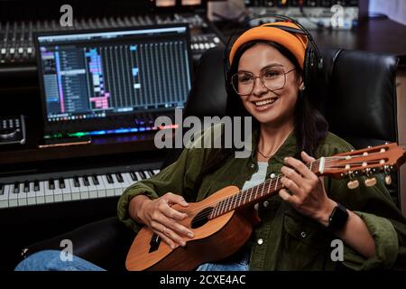 Ritratto di bella giovane brunetta, artista femminile seduto in studio di registrazione e giocando ukulele Foto Stock