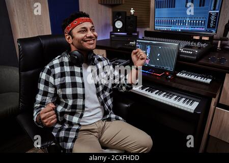 Giovane uomo, artista maschile che guarda felice con due pugni in aria mentre riesce a creare musica, seduto in studio di registrazione Foto Stock