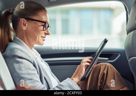 Lato di una donna di affari sorridente di successo che indossa occhiali utilizzando un tablet digitale, lavorando mentre si siede sul sedile posteriore in auto Foto Stock