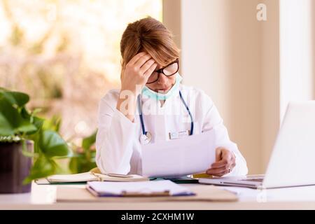 Foto di una dottoressa preoccupata che fa dei documenti mentre si siede alla scrivania dietro il suo laptop nell'ufficio del medico. Foto Stock