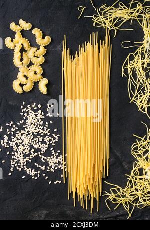 Assortimento di spaghetti di pasta secca, orzo, tagliatelle e creste di gallo su fondo testurizzato nero. Vista dall'alto Foto Stock