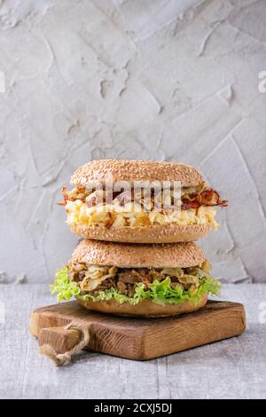 Pila di due panini con spezzatino di manzo, insalata fresca, pancetta, uova strapazzate e cipolla fritta servita sul piccolo tagliere di legno bianco su un tavolo di legno. Foto Stock