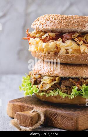 Pila di due panini con spezzatino di manzo, insalata fresca, pancetta, uova strapazzate e cipolla fritta servita sul piccolo tagliere di legno bianco su un tavolo di legno. Foto Stock