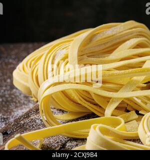 Raw uncooked homemade italian pasta tagliatelle with flour on old wood cutting board over dark wooden background. Close up. Square image Foto Stock