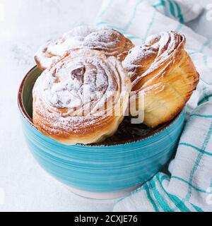 Moderni crostini di pasticceria, come croissant e muffin con zucchero in polvere, serviti in ciotola turchese con setaccio e asciugamano da cucina su sfondo di legno bianco Foto Stock