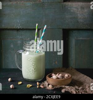 Yogurt da bere con pistacchi serviti in vasetto di vetro con tubetti da cocktail e ciotola di noci su vecchio tavolo di legno. Stile rustico scuro. Immagine quadrata Foto Stock