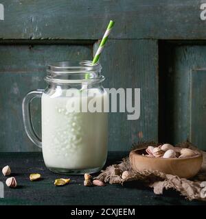 Yogurt da bere con pistacchi serviti in vasetto di vetro con tubetti da cocktail e ciotola di noci su vecchio tavolo di legno. Stile rustico scuro. Immagine quadrata Foto Stock