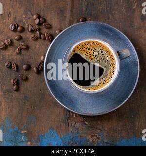 Tazza in ceramica blu di caffè nero caldo sul piatto, servita con chicchi di caffè su vecchio fondo testurizzato in legno. Vista dall'alto. Spazio di copia. Immagine quadrata Foto Stock