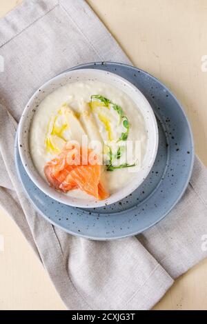 Ciotola di zuppa di crema di asparagi bianchi con germogli di piselli, salmone salato e pane tostato su tovagliolo grigio su sfondo bianco di legno. Disposizione piatta Foto Stock