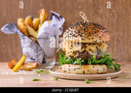 Hamburger fatti in casa con manzo, cipolla fritta e germogli di piselli, serviti su piatto di ceramica con patate fritte su fondo testurizzato di legno Foto Stock