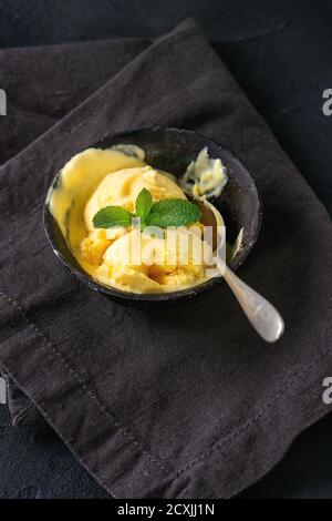 Gelato al mango fatto in casa con menta fresca in una ciotola di ferro vintage su un tovagliolo di tessuto scuro su sfondo nero testurizzato. Foto Stock