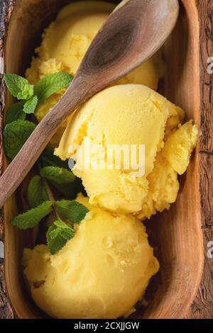 Gelato artigianale al mango con menta fresca in ciotola in legno d'oliva con cucchiaio di legno, servito su tovagliolo tessile bianco su fondo testurizzato in legno. Via superiore Foto Stock