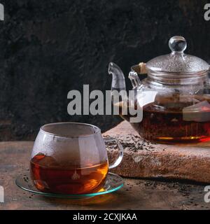 Tazza di vetro con piatto e teiera con tè nero caldo, in piedi su tavola di argilla su vecchio sfondo di legno. Con spazio per la copia. Immagine quadrata Foto Stock