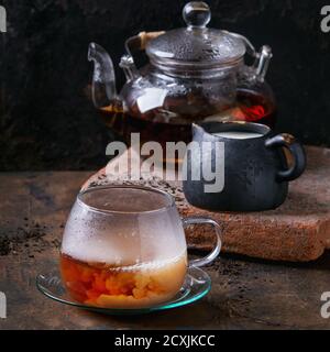 Tazza di vetro su piatto di tè nero con latte, teiera con tè nero caldo, in piedi su bordo di argilla con brocca di latte su vecchio sfondo di legno. Con copia s Foto Stock