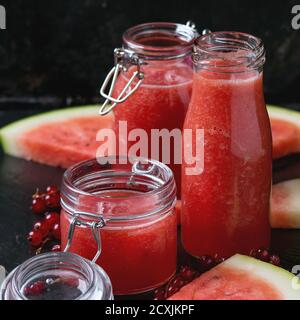 Vasetti di vetro e bottiglia di cocomero e frullato di ribes rosso, serviti con un pezzo di cocomero e frutti di bosco freschi su sfondo di ardesia nera. Con copia Foto Stock