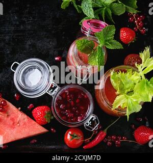 Varietà di frullati rossi di pomodoro, fragola e melograno in vasi di vetro, serve con menta, gambo di sedano e frutta fresca e verdure su blac Foto Stock