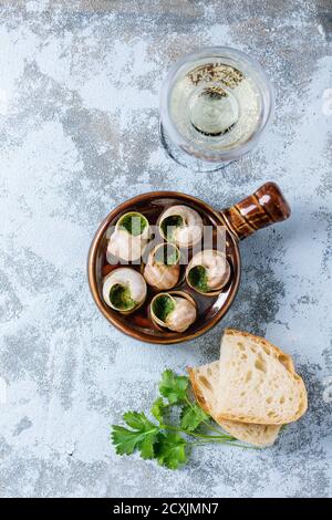 Escargots de Bourgogne - lumache con burro alle erbe, in tradizionale padella di ceramica con prezzemolo, pane e bicchiere di vino bianco su fondo testurizzato blu Foto Stock