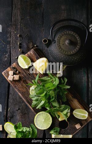 Ingredienti per tè verde ghiaccio lime, limone, menta, zucchero, tè verde e cubetti di ghiaccio su trivella di legno con bicchiere di coctail e teiera di ferro nero sopra Foto Stock