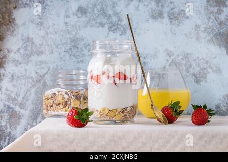 Sana colazione muesli, fragole e yogurt con il frullato di mango in vetro in vasetti di vetro. Servita con fragole fresche sulla tovaglia bianca con textu Foto Stock