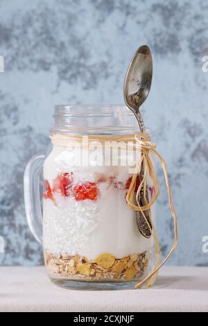 Sana colazione muesli, fragole e yogurt in vaso di vetro mason con cucchiaio. Yogurt servito su tovaglie bianche di tessuto con parete testurizzata sul retro Foto Stock