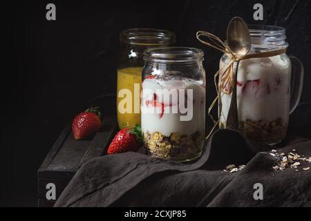 Sana colazione muesli, fragole e yogurt con il frullato di mango in vetro in vasetti di vetro. Servita sul tagliere di legno con fragole fresche. Dark Foto Stock
