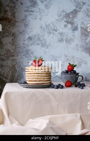 Prima colazione con frittelle fresche e mirtilli in piatto di ceramica blu e in piccola tazza, servita su tovaglia di lino bianco con tex blu Foto Stock