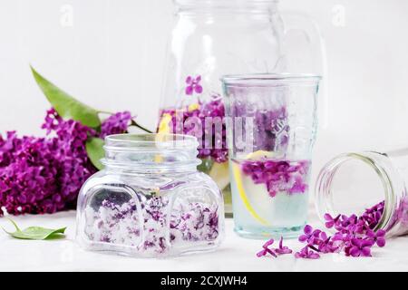 Vaso di vetro di fiori lilla in zucchero, vetro e caraffa di acqua lilla con limone e ramo di lilla fresco su tovaglia di lino bianco. Foto Stock