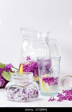 Vaso di vetro di fiori lilla in zucchero, vetro e caraffa di acqua lilla con limone e ramo di lilla fresco su tovaglia di lino bianco. Foto Stock