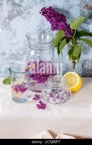 Il vasetto di vetro di lilla fiori in zucchero, il vetro e la brocca di lillà Acqua con limone e il ramo di lilla fresca sulla biancheria da letto bianca tovaglia con textur blu Foto Stock