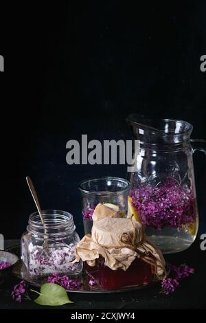 Vasetto di vetro di sciroppo di lilla fatto in casa, vasetto di vetro di fiori di lilla zuccherati e caraffa di vetro di limonata lilla acqua su tovaglia nero su nero. Oscura r Foto Stock