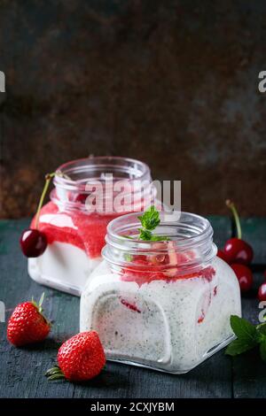 Due vasetti di vetro quadrati con yogurt fatto in casa con menta, purea di fragole e ciliegia, serviti con frutti di bosco freschi su sfondo di legno scuro. Con spazio f Foto Stock