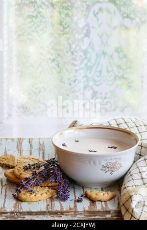Biscotti alla lavanda e ciotola di latte aromatico, serviti con asciugamano da cucina su vecchio tavolo di legno con finestra sullo sfondo. Prima colazione in stile rustico, natura Foto Stock