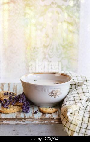 Biscotti alla lavanda e ciotola di latte aromatico, serviti con asciugamano da cucina su vecchio tavolo di legno con finestra sullo sfondo. Prima colazione in stile rustico, natura Foto Stock