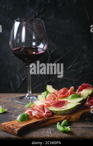 Melone affettato con prosciutto e foglie di basilico, servito su tagliere di legno d'oliva con un bicchiere di vino rosso su vecchio fondo testurizzato di legno. Con spazio per Foto Stock