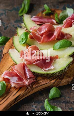 Melone affettato con prosciutto e foglie di basilico, servito su tagliere di legno di oliva su vecchio sfondo testurizzato di legno. Foto Stock