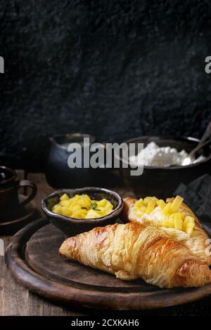 La prima colazione con due cornetti, burro, formaggio, caffè e fette di mango fruit, servita su legno tagliere sopra il vecchio sfondo di legno. Chiudere u Foto Stock