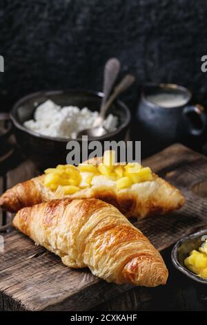 La prima colazione con due cornetti, burro, formaggio, caffè e fette di mango fruit, servita su legno tagliere sopra il vecchio sfondo di legno. Chiudere u Foto Stock