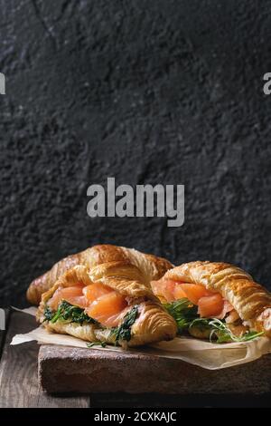 Due croissant con affumicati Salmoni salati, spinaci e rucola servita sulla carta da forno di terracotta oltre il bordo scuro legno vecchio sfondo. Spazio di copia Foto Stock