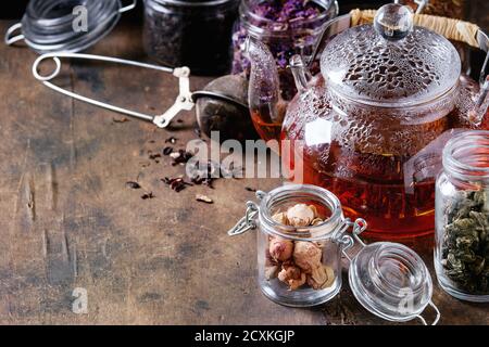 Varietà di foglie di tè nero, verde e a base di erbe secche in vasi di vetro con filtro vintage e teiera di tè caldo su vecchio sfondo di legno scuro. Primo piano, Foto Stock