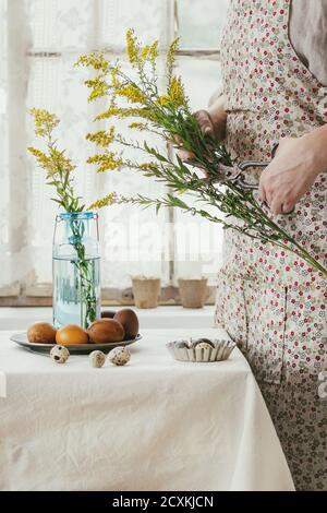 Preparandosi per Pasqua. Le mani femminili tagliano con le vecchie forbici fiori gialli vicino tavolo tovaglie bianco decorato pollo marrone e quaglia di colore pasqua Foto Stock