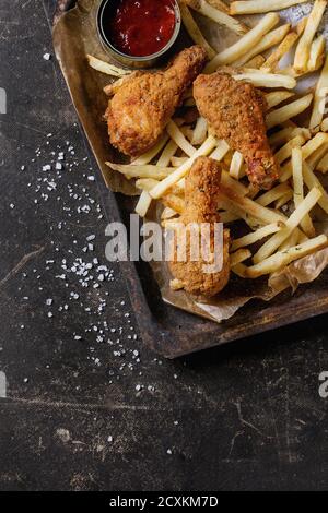 Il fast food fritto croccante di cosce di pollo e patatine fritte di patate con sale e ketchup salsa servita sulla carta da forno nel vecchio forno arrugginito il vassoio su testo scuro Foto Stock