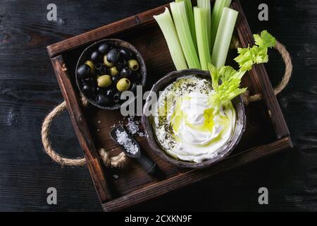 Sedano fresco con yogurt e olio d'oliva tuffo nella ciotola in ceramica, servita con sale marino e nero, olive verdi sul vassoio di legno su legno nero backgrou bruciato Foto Stock