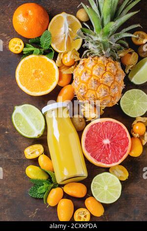 Varietà di agrumi interi e affettati ananas, pompelmo, limone, lime, kumquat, clementina e fisalis con menta e bottiglia di succo al buio Foto Stock