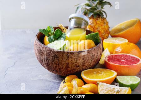 Varietà di agrumi interi e affettati ananas, pompelmo, limone, lime, kumquat, clementina e fisalis con menta e bottiglia di succo nel recipiente o Foto Stock