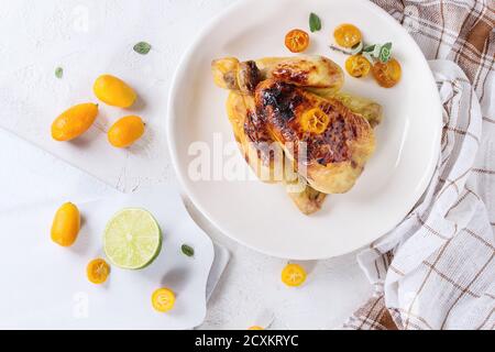 Tutta la cottura di mini pollo arrosto con kumquat caramellati servita su piatto bianco con asciugamano da cucina, taglieri e agrumi freschi sopra chi Foto Stock