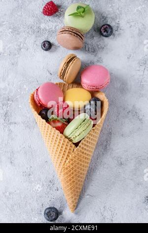 Varietà di colorati macaron dolci francesi con diversi macaron ripieni serviti nel cono del waffle con menta e frutti di bosco freschi trama grigia Foto Stock